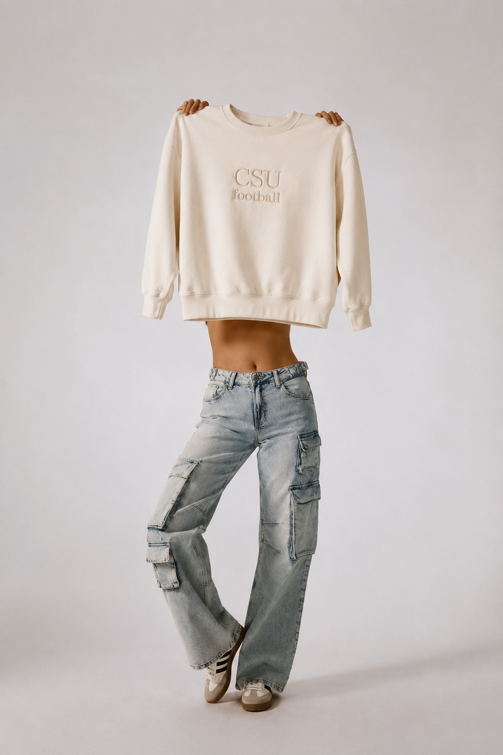 A model holds up a cream-colored crewneck sweatshirt with 'CSU football' printed in gold on the chest, styled with light-wash distressed jeans and sneakers against a light gray studio background.