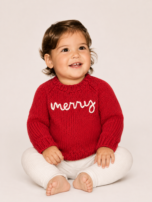 A smiling toddler sitting cross-legged on a light beige studio background, wearing a red knit sweater with 'merry' written across the chest in white script and white pants.