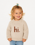 A young toddler girl with blonde hair in a small bun wearing a cream-colored chunky knit sweater with the word 'hi.' knitted in brown across the chest, paired with light blue jeans, against a plain white background.