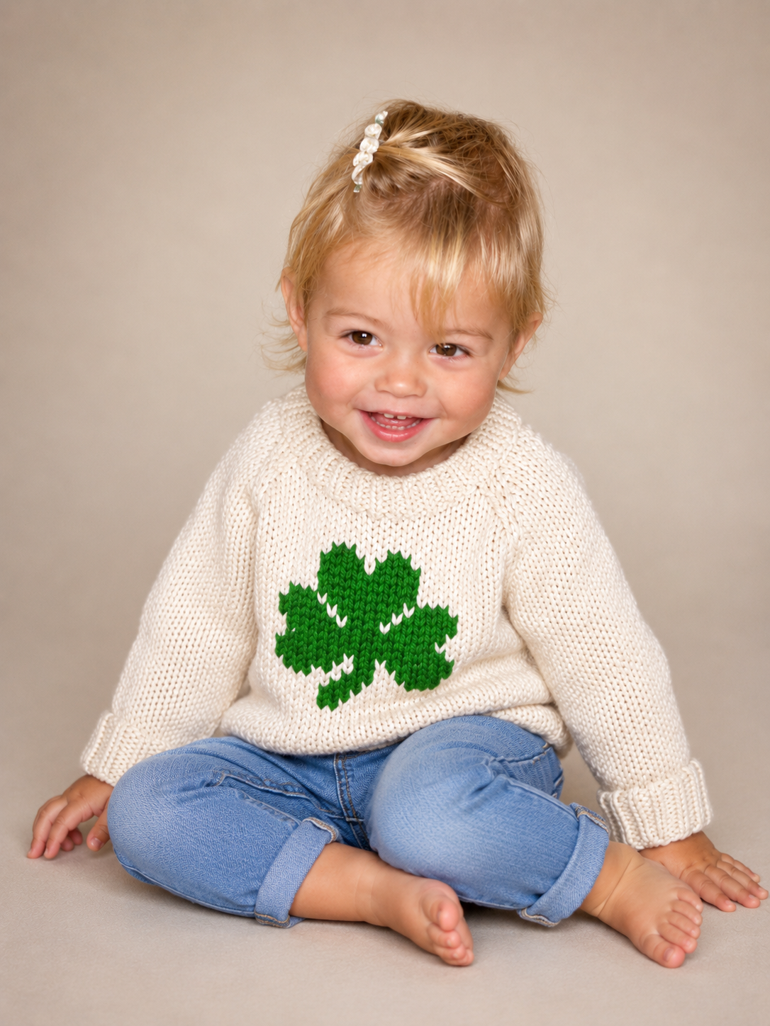 A smiling toddler sitting cross-legged on a beige studio floor, wearing a cream knit sweater with a green shamrock on the front and blue jeans.