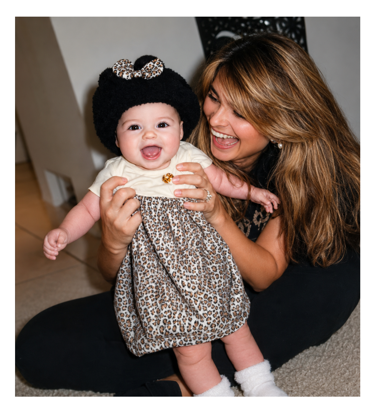 A baby wearing a black knit hat with a leopard-print bow and a leopard-print dress, held up by a smiling woman in a black outfit.