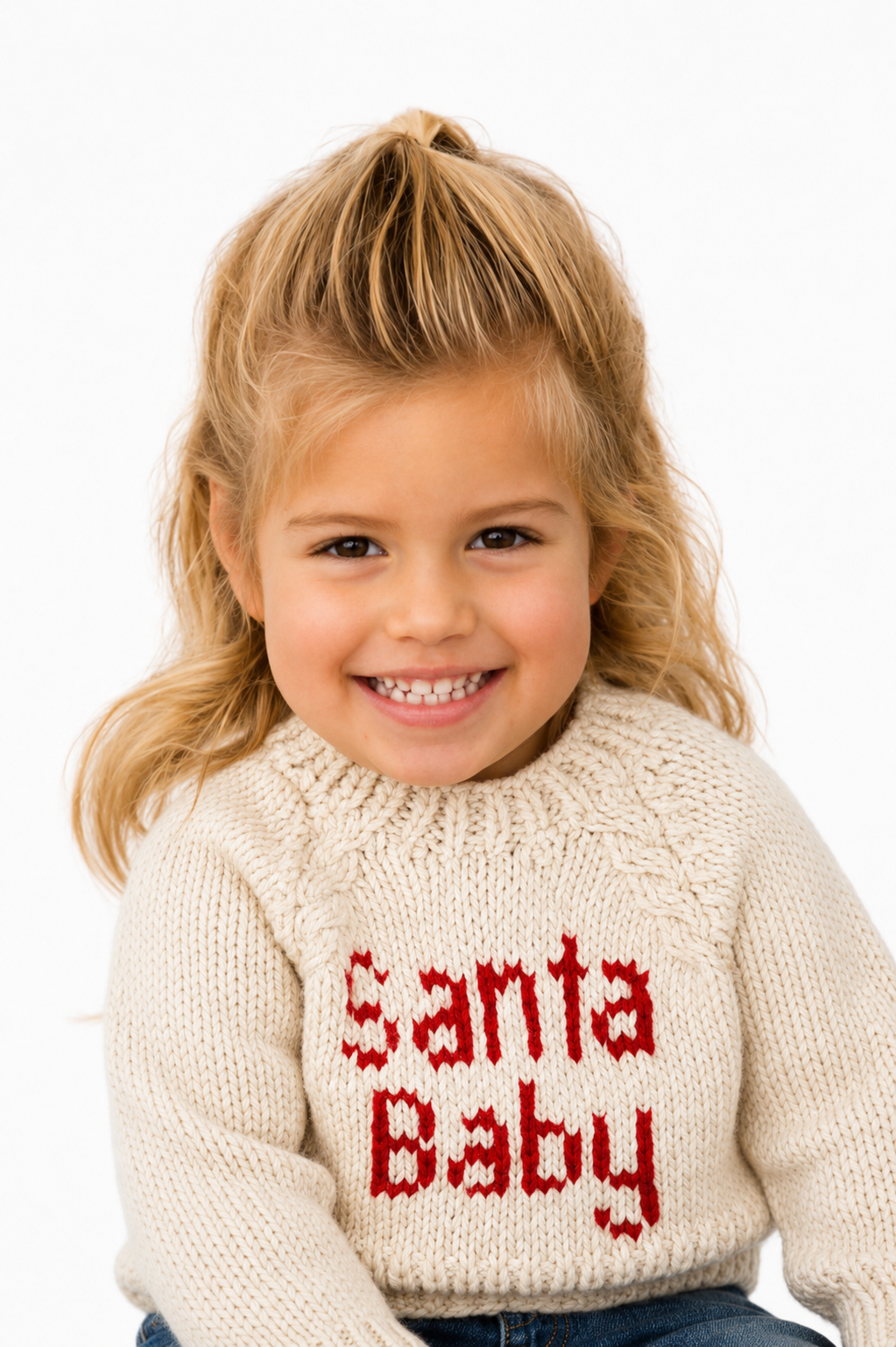A young girl with blonde hair wearing a cream knit sweater with 'Santa Baby' written in red across the chest, photographed against a plain white background.