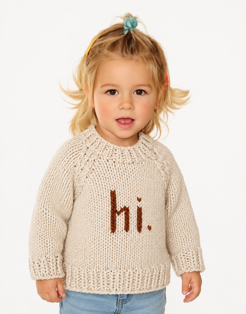A toddler girl with blonde hair in a small blue bow wears a cream-colored knit sweater with the word 'hi.' in brown lettering on the front, paired with light blue jeans, against a plain white background.