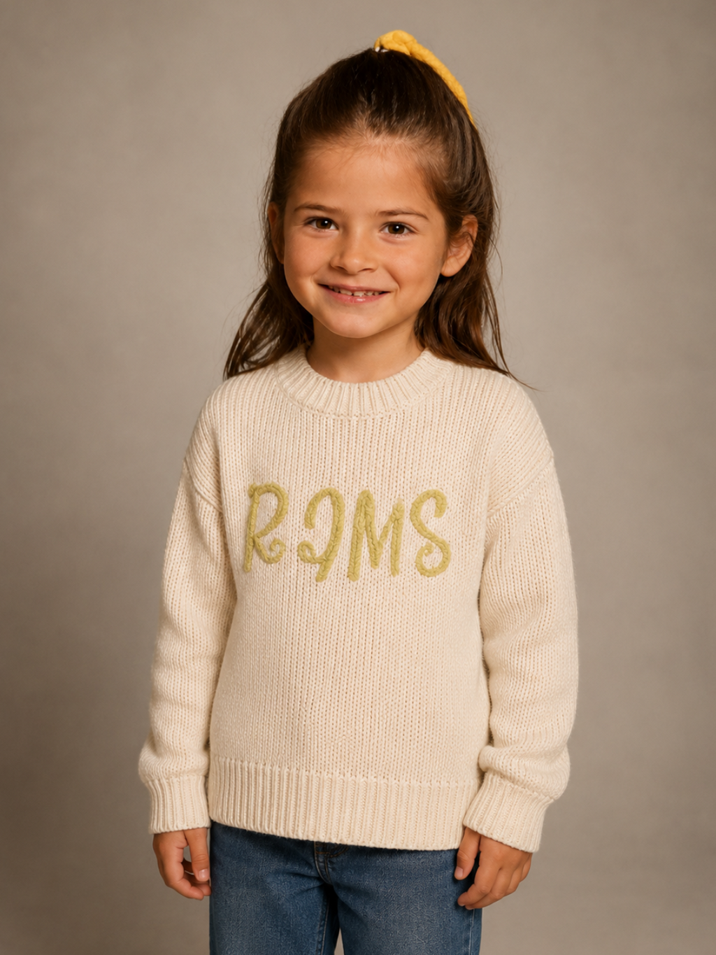 A young girl with her hair in a yellow bow stands against a neutral gray studio background, wearing a cream ribbed knit sweater with 'RIMS' in gold glitter lettering across the chest, paired with blue jeans.