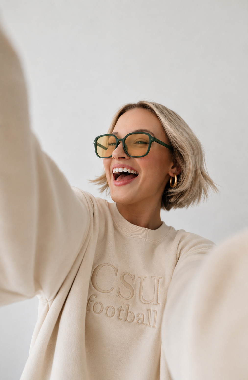 A woman with short blonde hair wearing green-framed glasses and a cream sweatshirt with 'CSU football' embroidered on the front, taking a selfie against a plain light background.