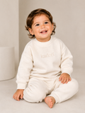 A smiling toddler sitting cross-legged on a light beige studio floor, wearing a matching cream sweatshirt and sweatpants set. The sweatshirt features the brand name 'kadori' printed in a subtle, tonal font on the chest.