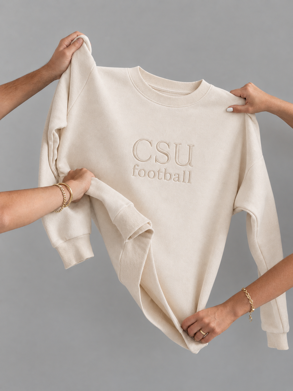 A cream-colored crewneck sweatshirt with 'CSU football' embroidered on the chest, held up by multiple hands against a solid gray background.