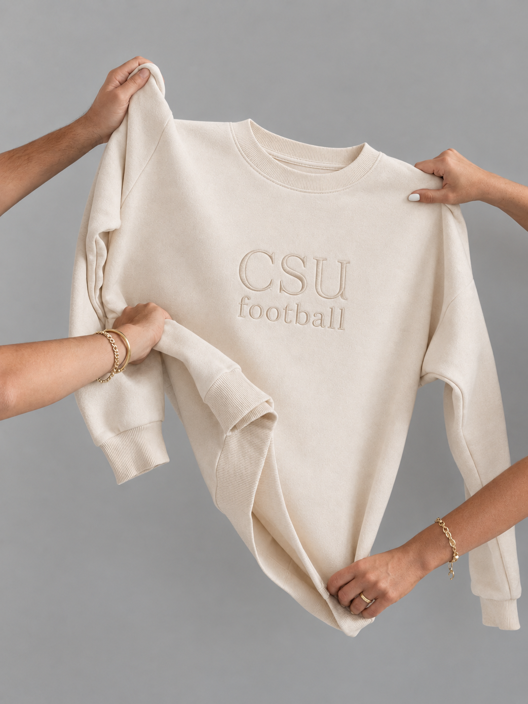 A cream-colored crewneck sweatshirt with 'CSU football' embroidered on the chest, held up by multiple hands against a solid gray background.