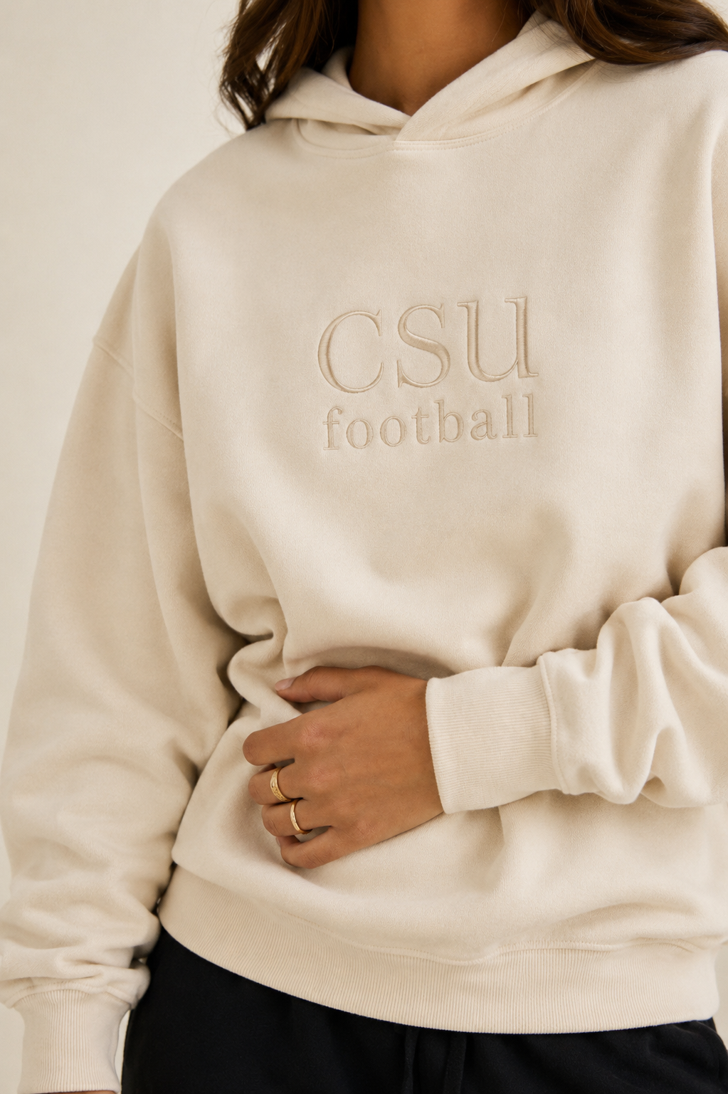 A person wearing a cream-colored hoodie with 'CSU football' embroidered on the chest, paired with black pants, against a plain light background.
