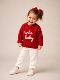 A toddler girl wearing a red knit sweater with 'Santa Baby' written in white script, paired with white pants and white sneakers, standing against a neutral beige background.