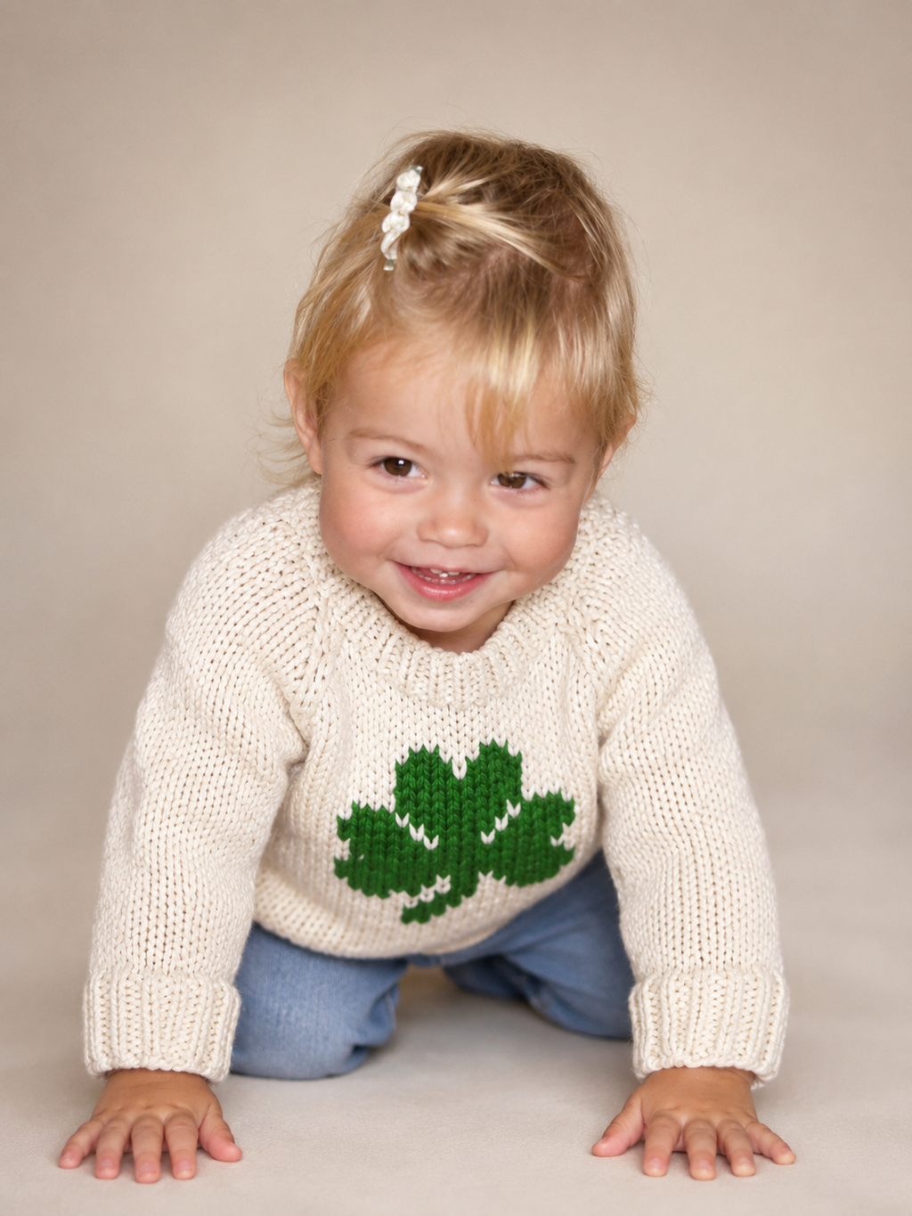 A toddler girl crawling on a beige studio background, wearing a cream knit sweater with a green shamrock on the front and blue pants.