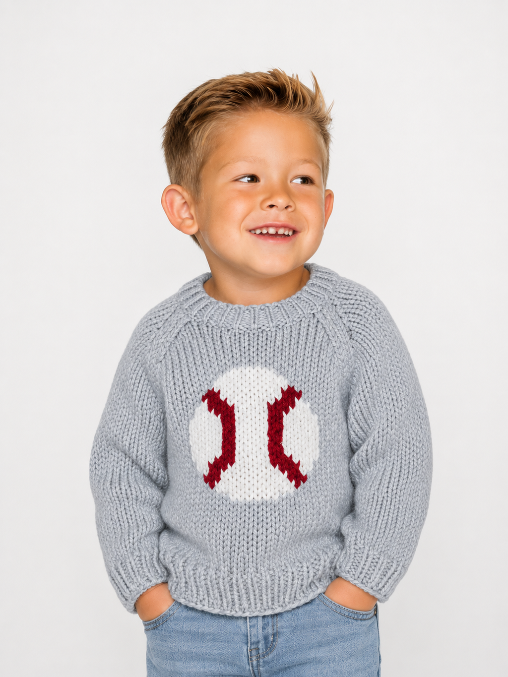 A young boy smiling and looking to the side, wearing a light gray knit sweater with a white baseball motif and red stitching details on the chest, paired with blue jeans, against a plain light gray background.