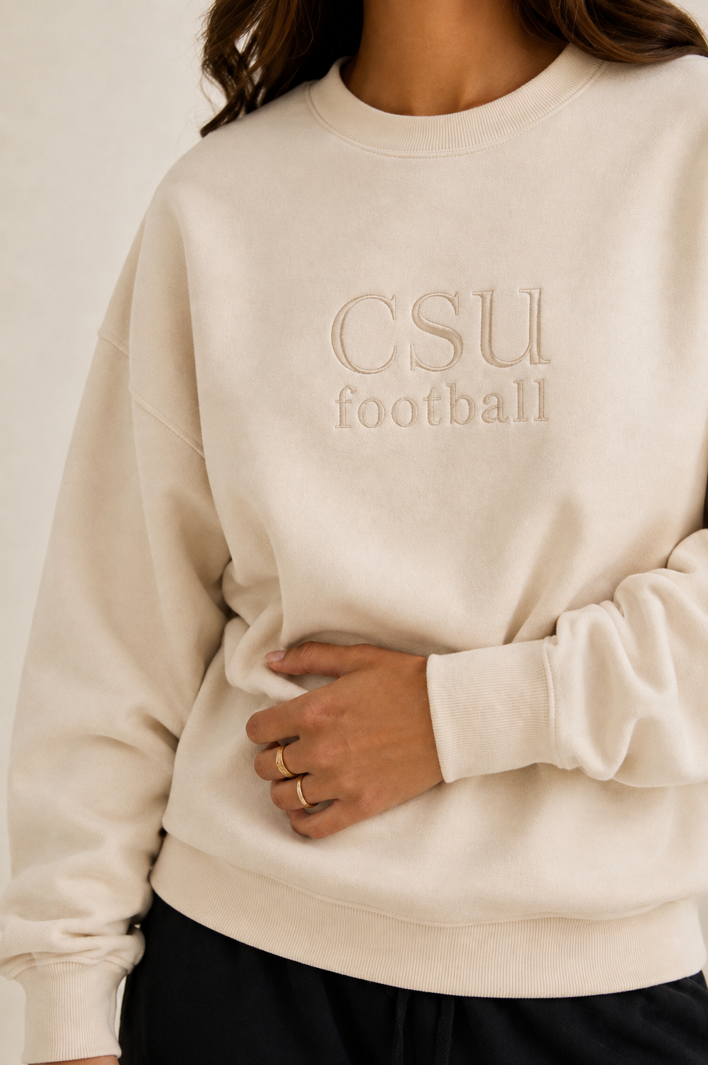 A close-up of a person wearing a cream-colored crewneck sweatshirt with 'CSU football' embroidered on the chest, paired with black pants against a neutral background.