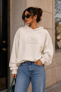 A woman wearing an off-white CSU football hoodie and blue jeans stands outdoors against a beige stone building wall.
