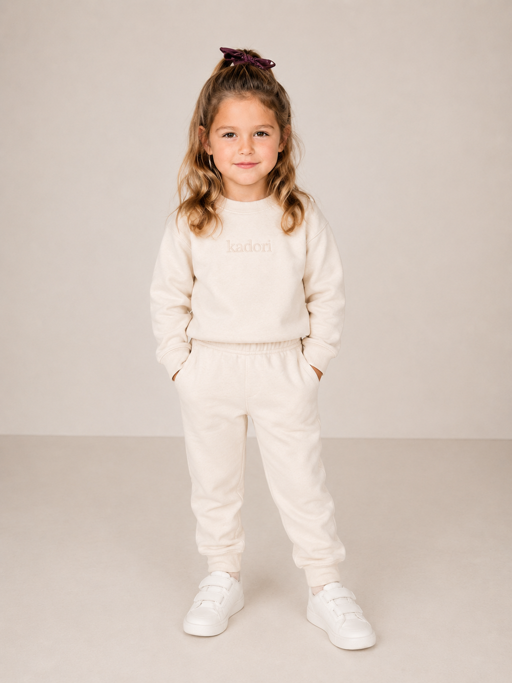 A young girl with blonde hair in a high ponytail wearing a cream-colored sweatshirt and matching sweatpants with white sneakers, standing against a neutral beige studio background.