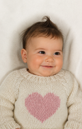 A smiling baby lying on a white blanket, wearing a cream knit sweater with a pink heart on the front.