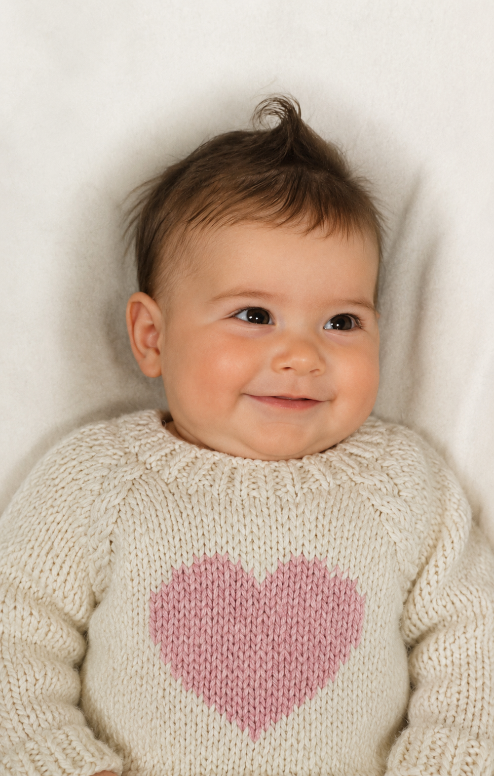A smiling baby lying on a white blanket, wearing a cream knit sweater with a pink heart on the front.