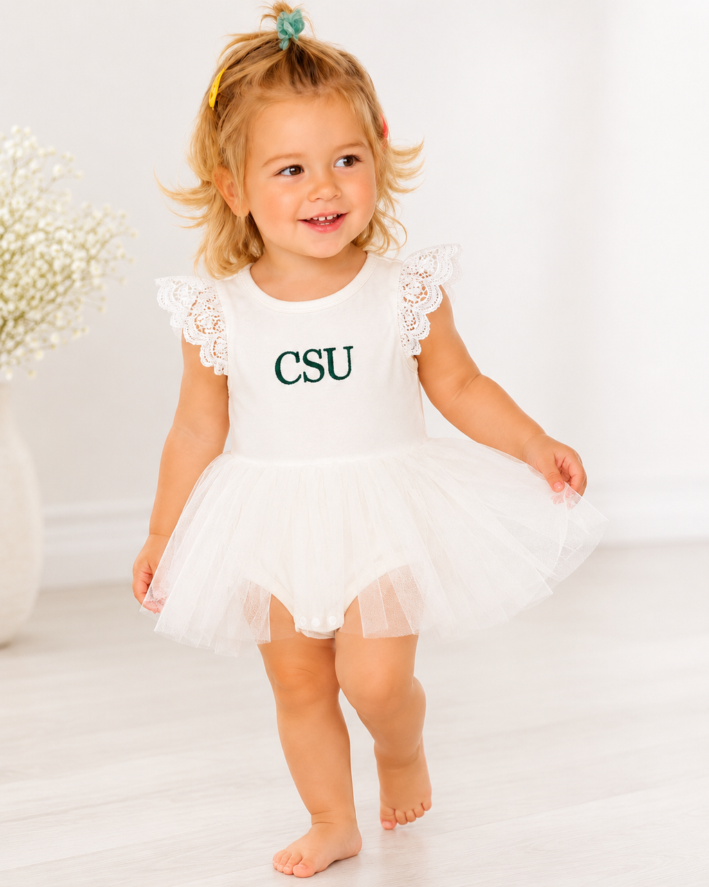 A toddler girl walking barefoot in a white sleeveless tutu dress with lace flutter sleeves and 'CSU' printed in green on the front, set against a bright white indoor background.