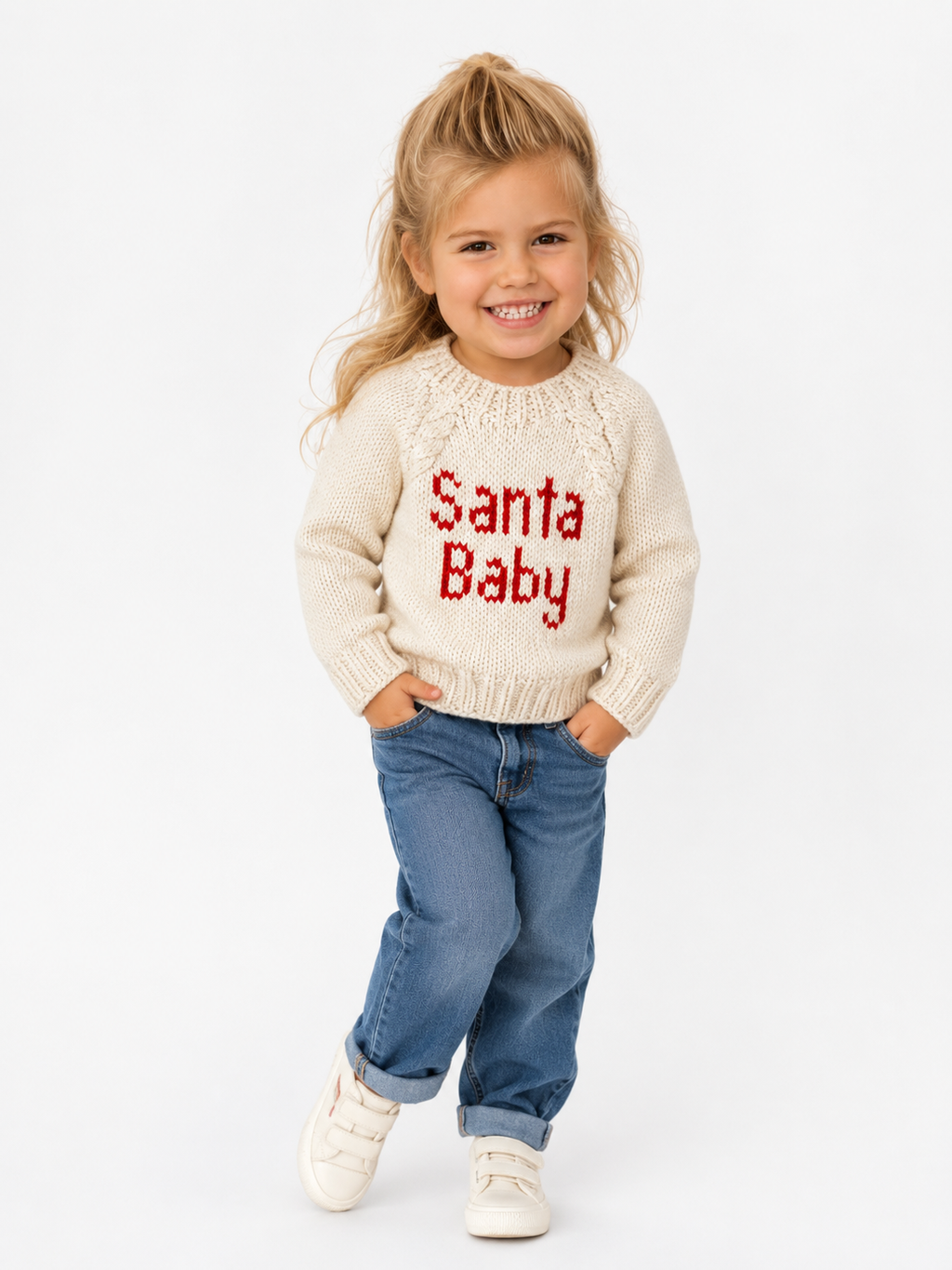 A young girl with blonde hair stands against a white studio background wearing a cream knit sweater with 'Santa Baby' written in red across the chest, blue jeans, and white sneakers.