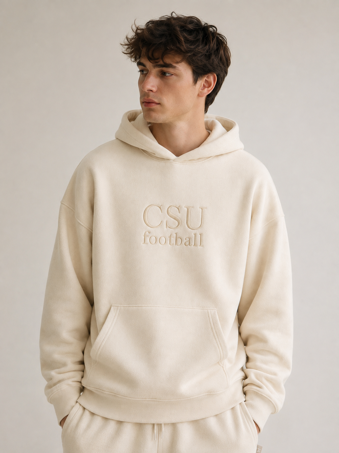 A man wearing a cream-colored hoodie with 'CSU football' embroidered on the chest, paired with matching sweatpants, set against a plain light gray studio background.