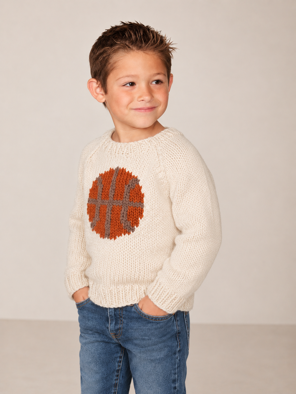 A young boy wearing a cream-colored knit sweater with a large orange and gray basketball graphic on the chest, paired with blue jeans, standing against a plain light beige background.