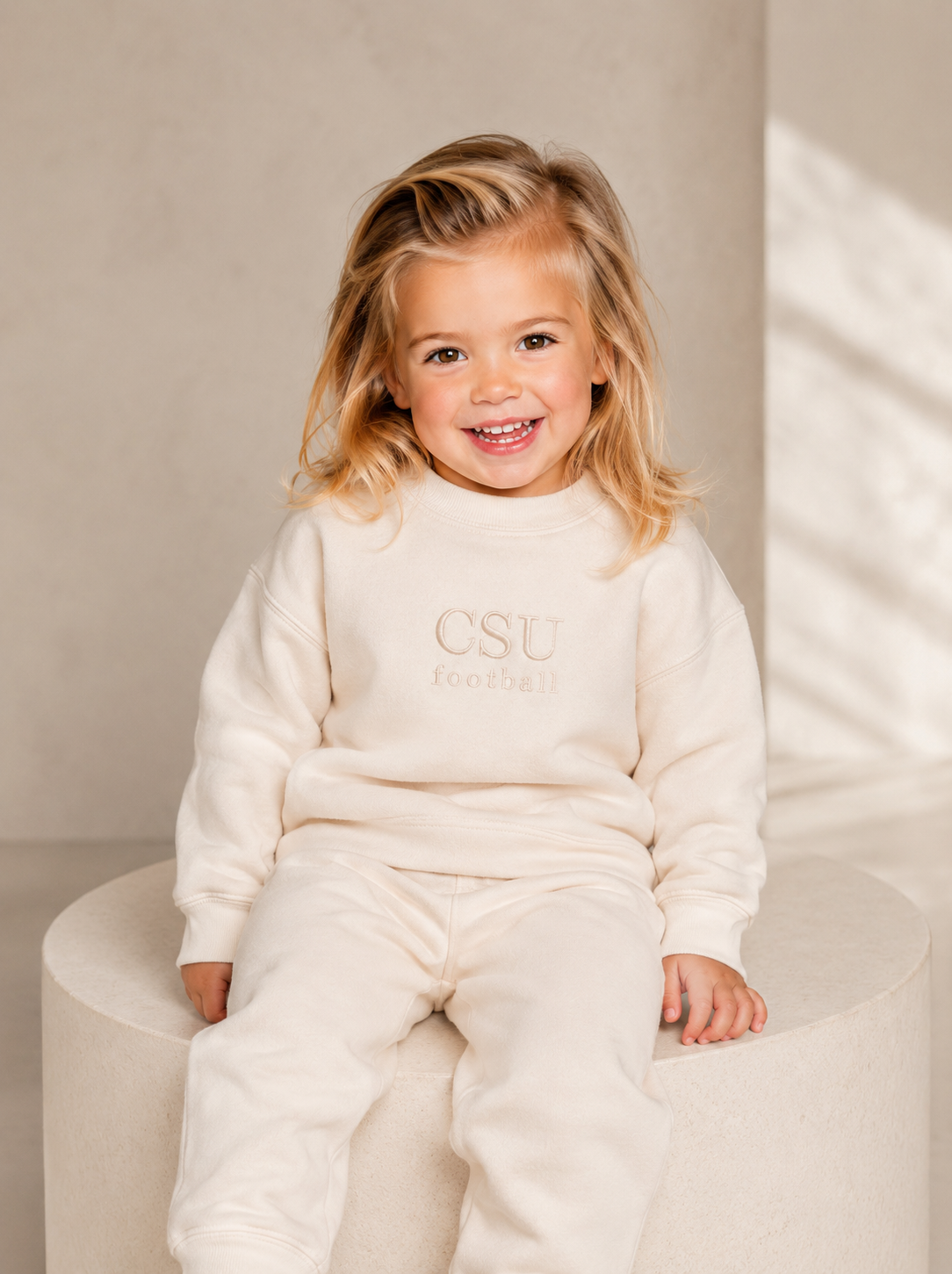 A young girl with blonde hair sits smiling on a round beige pedestal, wearing a cream-colored two-piece sweatshirt and jogger set with 'CSU Santa Barbara Football' printed on the chest.