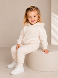 A young girl with blonde hair sits smiling on a round beige pedestal, wearing a cream-colored matching hoodie and jogger sweatpants set with white sneakers. The hoodie features a subtle 'CSU FOOTBALL' logo on the chest.