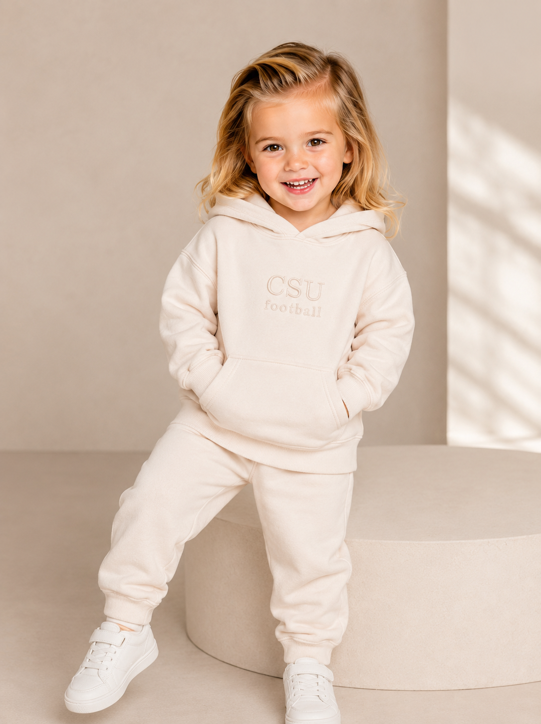 A young girl with blonde hair is smiling and posing in a matching cream-colored two-piece tracksuit with 'CSU football' printed on the chest, paired with white sneakers, set against a minimalist beige studio background.