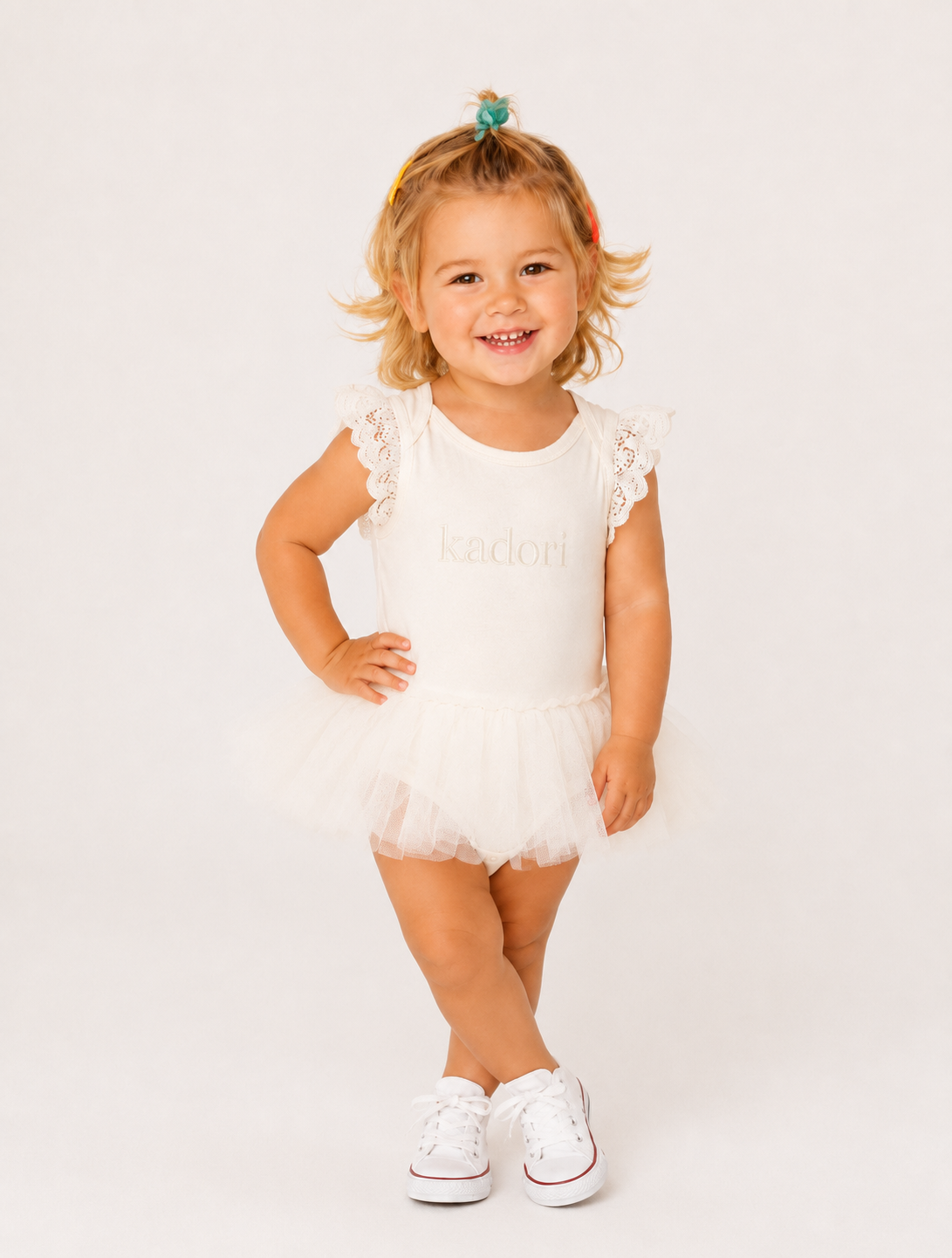A toddler girl with blonde hair wearing a white tutu dress with lace ruffle sleeves and white sneakers, standing against a plain white studio background.