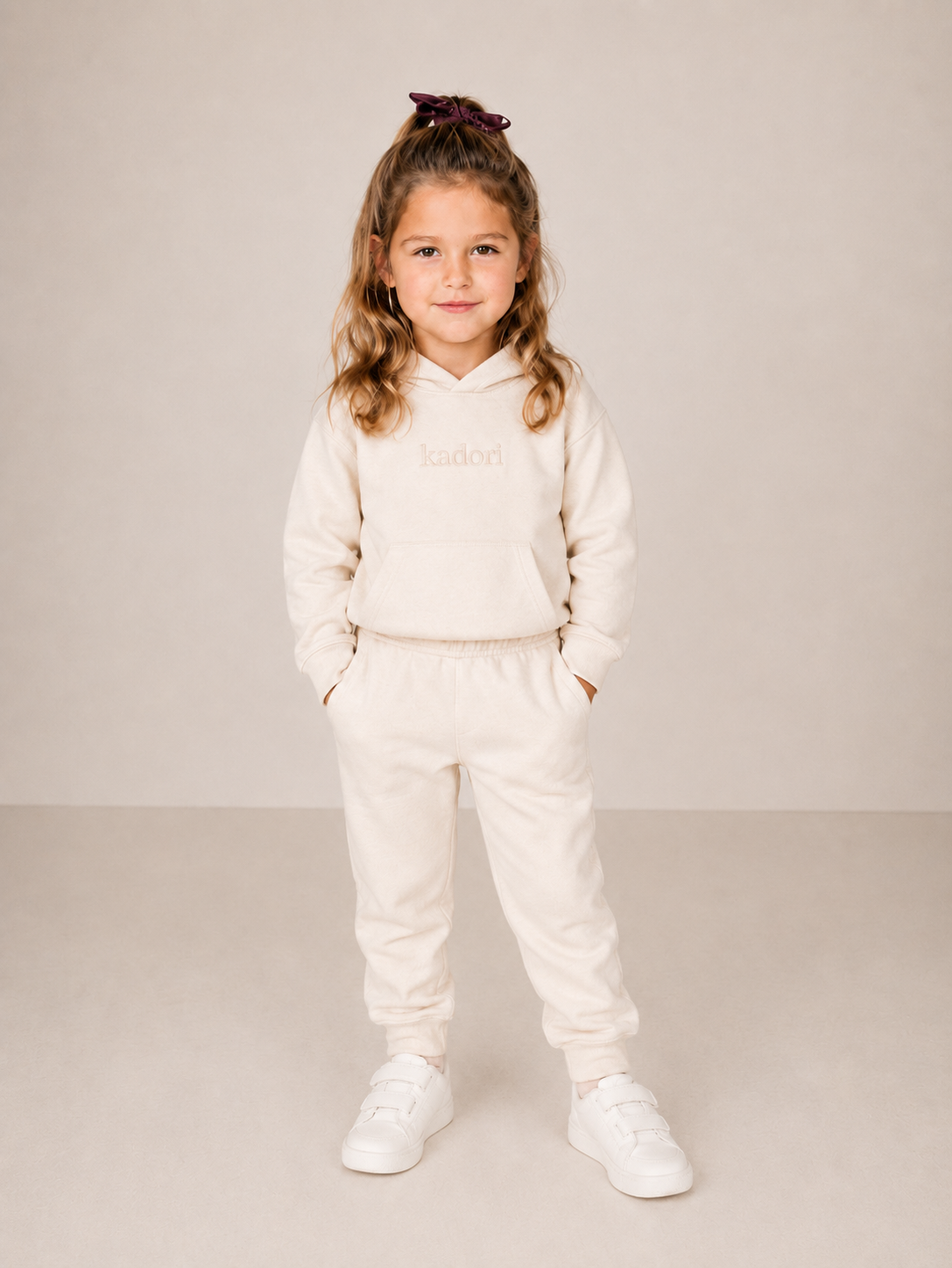 A young girl with light brown hair in a high ponytail wearing a cream-colored two-piece tracksuit with a small embroidered logo on the chest, paired with white sneakers, standing against a neutral beige studio background.