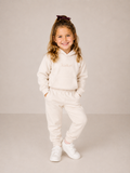 A young girl with blonde hair in a high ponytail wearing a cream-colored two-piece tracksuit with the brand name 'Laduri' printed on the chest, paired with white sneakers, standing against a neutral beige studio background.