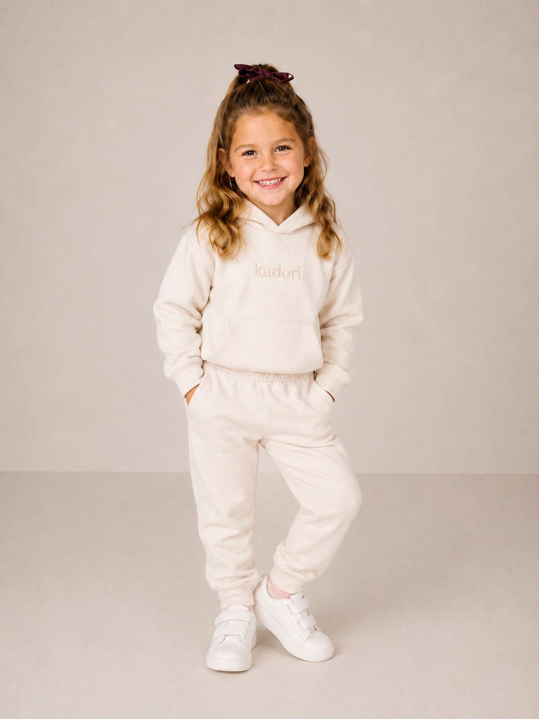 A young girl with blonde hair in a high ponytail wearing a cream-colored two-piece tracksuit with the brand name 'Laduri' printed on the chest, paired with white sneakers, standing against a neutral beige studio background.
