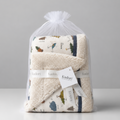 A folded baby blanket or towel set wrapped in a clear plastic bag with a white drawstring, featuring a cream base with a woodland print and a white ribbon label that reads Kindori.