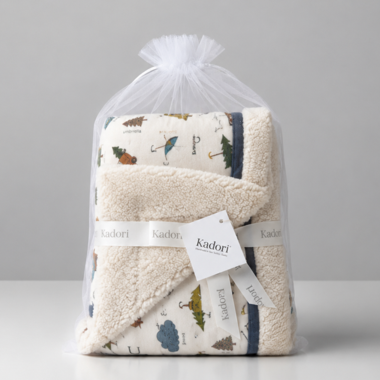 A folded baby blanket or towel set wrapped in a clear plastic bag with a white drawstring, featuring a cream base with a woodland print and a white ribbon label that reads Kindori.