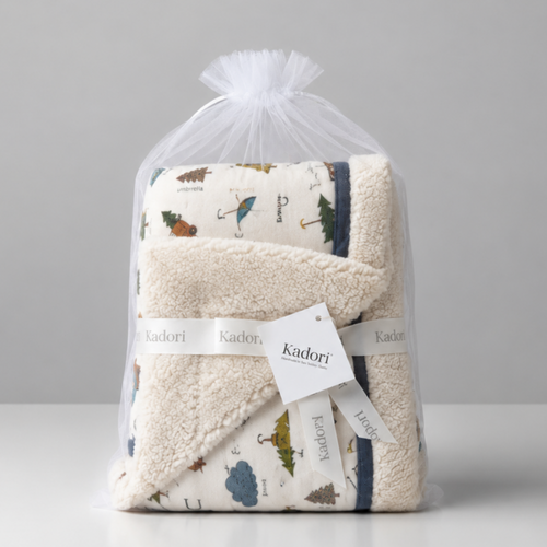 A folded baby blanket or towel set wrapped in a clear plastic bag with a white drawstring, featuring a cream base with a woodland print and a white ribbon label that reads Kindori.