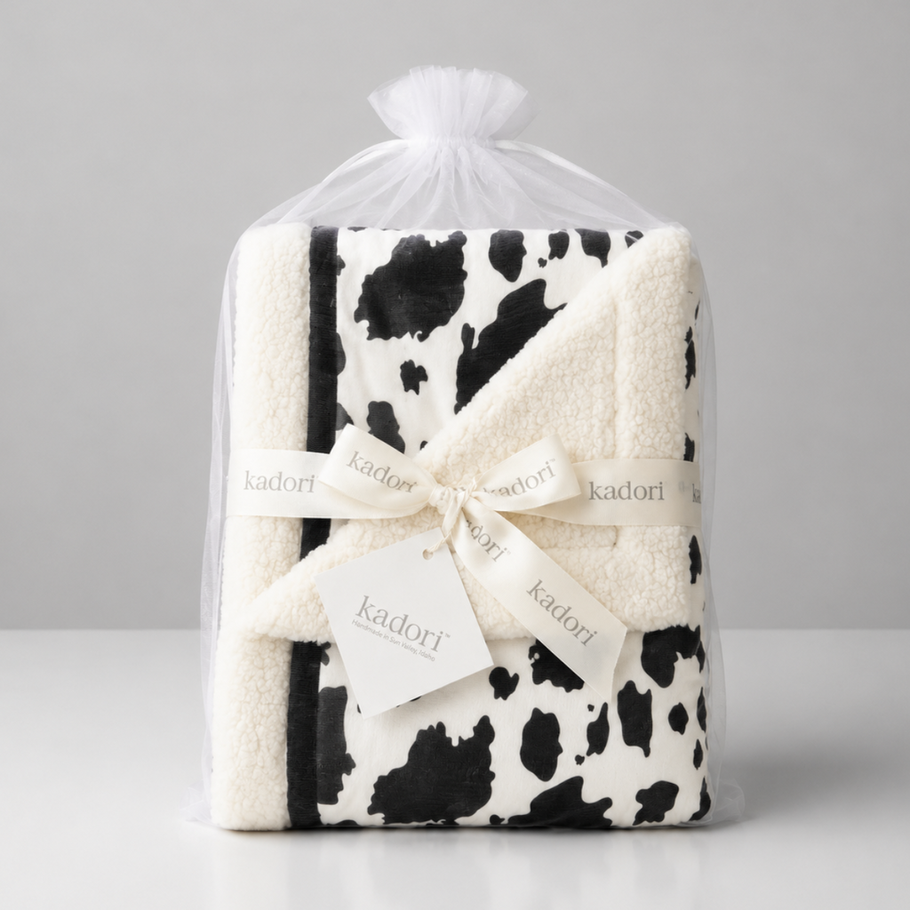 A folded plush blanket with a black and white cow print pattern, wrapped in a clear plastic bag and tied with a cream ribbon printed with the brand name 'kadori'.