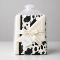 A folded plush blanket with a black and white cow print pattern, wrapped in a clear plastic bag and tied with a cream ribbon printed with the brand name 'kadori'.