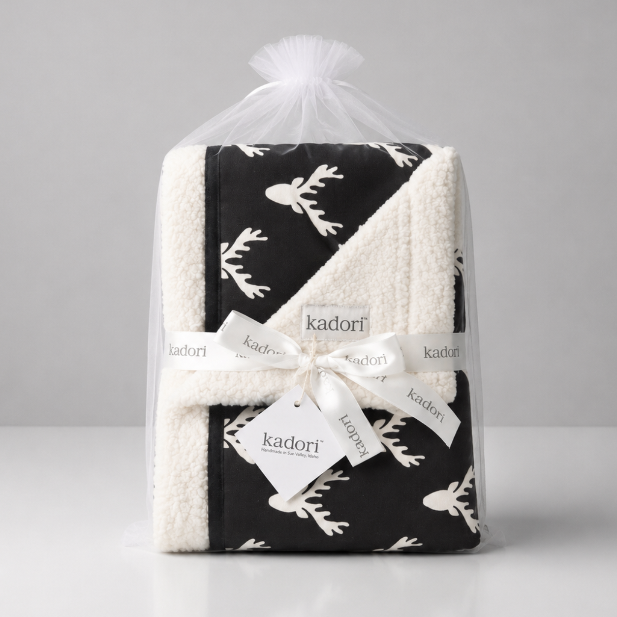 A folded baby blanket with a black and white reindeer pattern, wrapped in a clear cellophane bag and tied with a white ribbon printed with the brand name 'kadorf'.