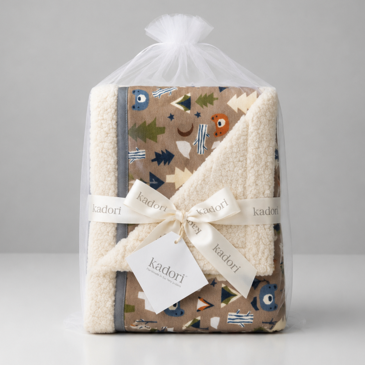 A folded baby blanket set by Kadori wrapped in a clear tulle bag, tied with a cream ribbon, and presented on a light gray background. The blanket features a soft cream sherpa fleece side and a patterned side with a brown background, blue owl illustrations, and green and orange tree accents.