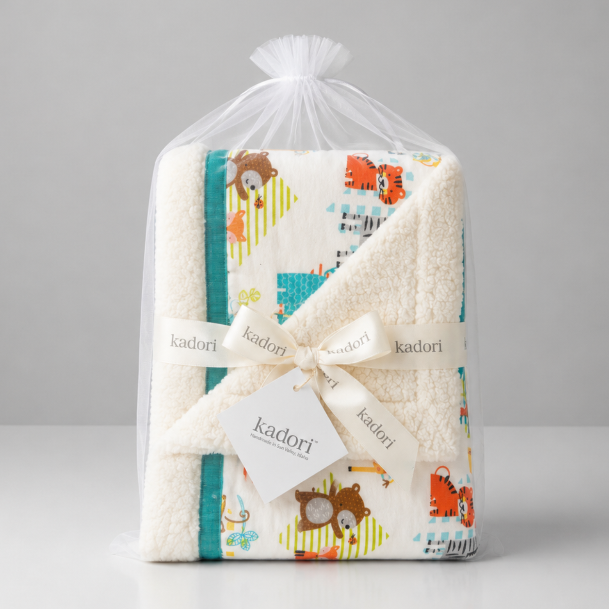 A folded baby blanket wrapped in a clear plastic bag, tied with a cream ribbon and tag. The blanket has an off-white sherpa lining and a printed front panel featuring colorful cartoon animals and patterns on a light background.