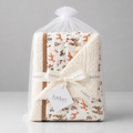 A packaged Kadori baby blanket set wrapped in a clear plastic bag with a white pom-pom, tied with a white ribbon, and featuring a woodland print on a cream background. The set includes a fleece blanket and a plush insert, both with a brown border.