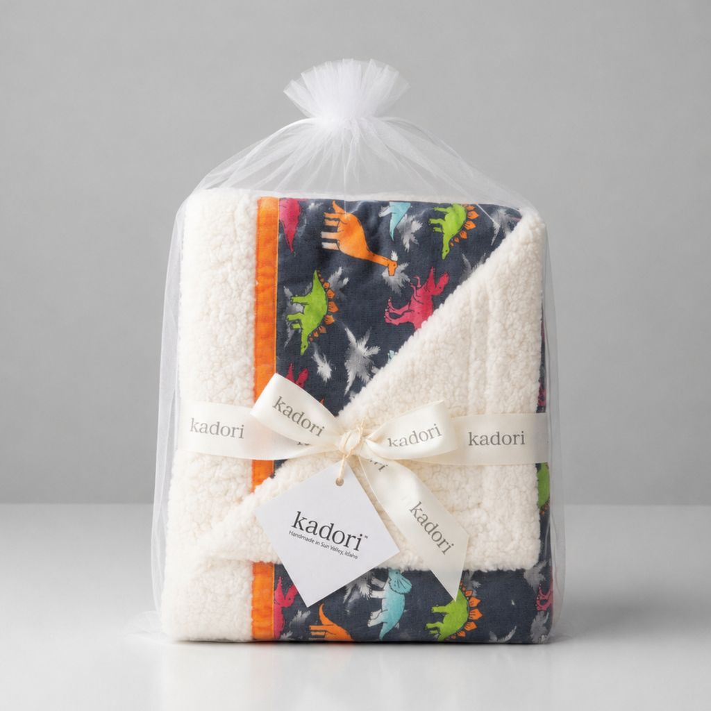 A packaged baby blanket from Kadori, wrapped in a clear plastic bag with a white ribbon and tag. The blanket features a white sherpa fleece reverse side and a dark navy print side with colorful dinosaur illustrations and orange stripes.