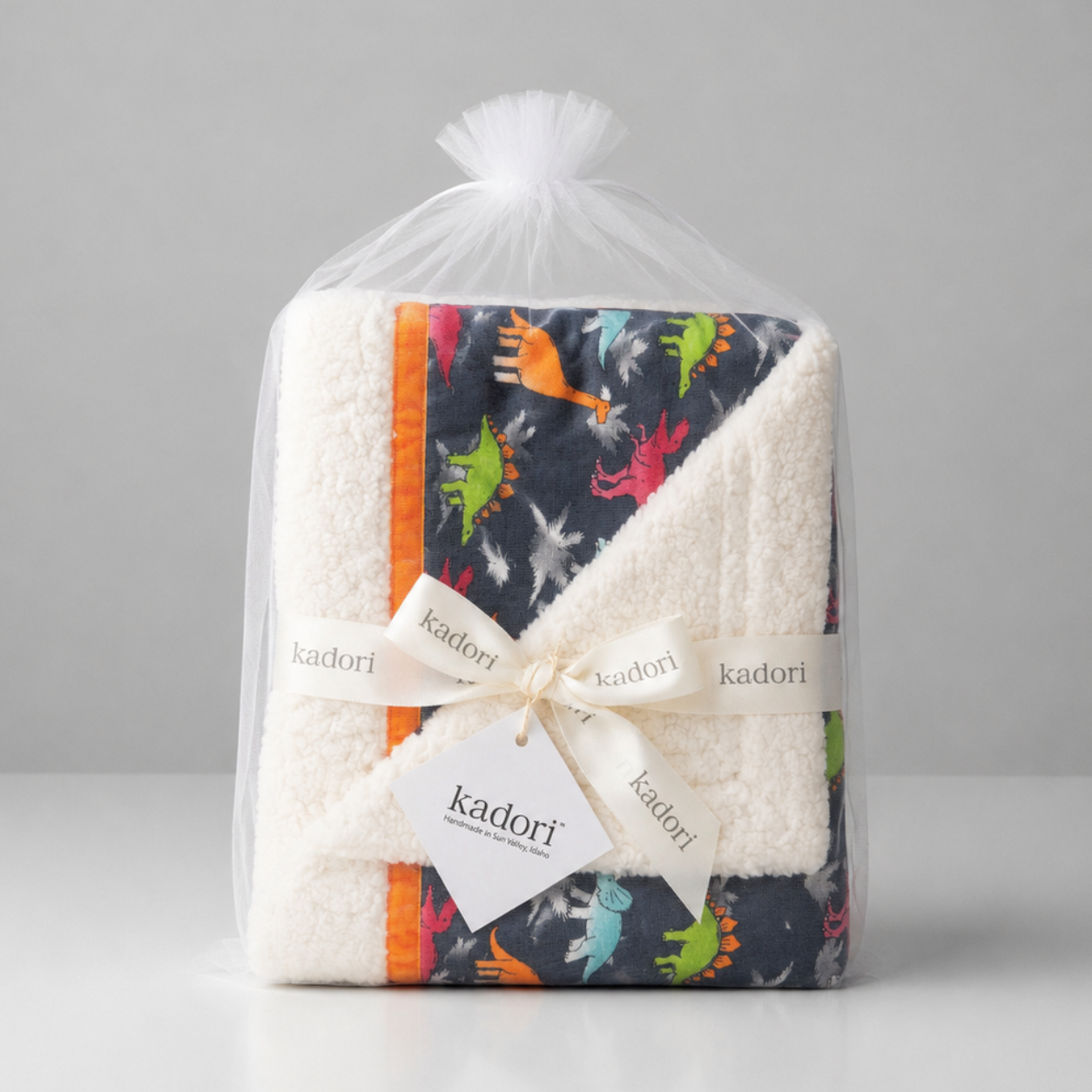 A packaged baby blanket from Kadori, wrapped in a clear plastic bag with a white ribbon and tag. The blanket features a white sherpa fleece reverse side and a dark navy print side with colorful dinosaur illustrations and orange stripes.