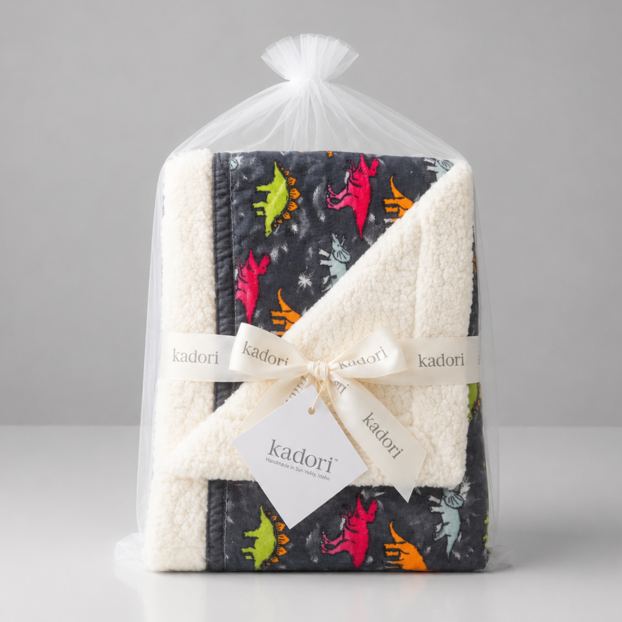A folded Kadori baby blanket set wrapped in a clear cellophane bag and tied with a white ribbon, featuring a dark gray printed fabric with colorful dinosaur silhouettes and a cream sherpa lining.