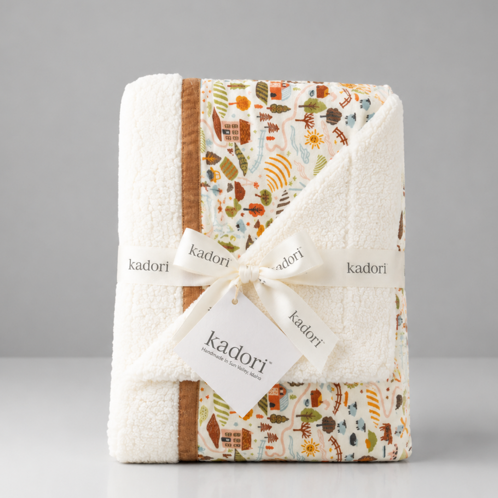 A folded Kadori baby blanket with a cream sherpa border and a multicolor autumn-themed patchwork pattern, tied with a white ribbon and tag.