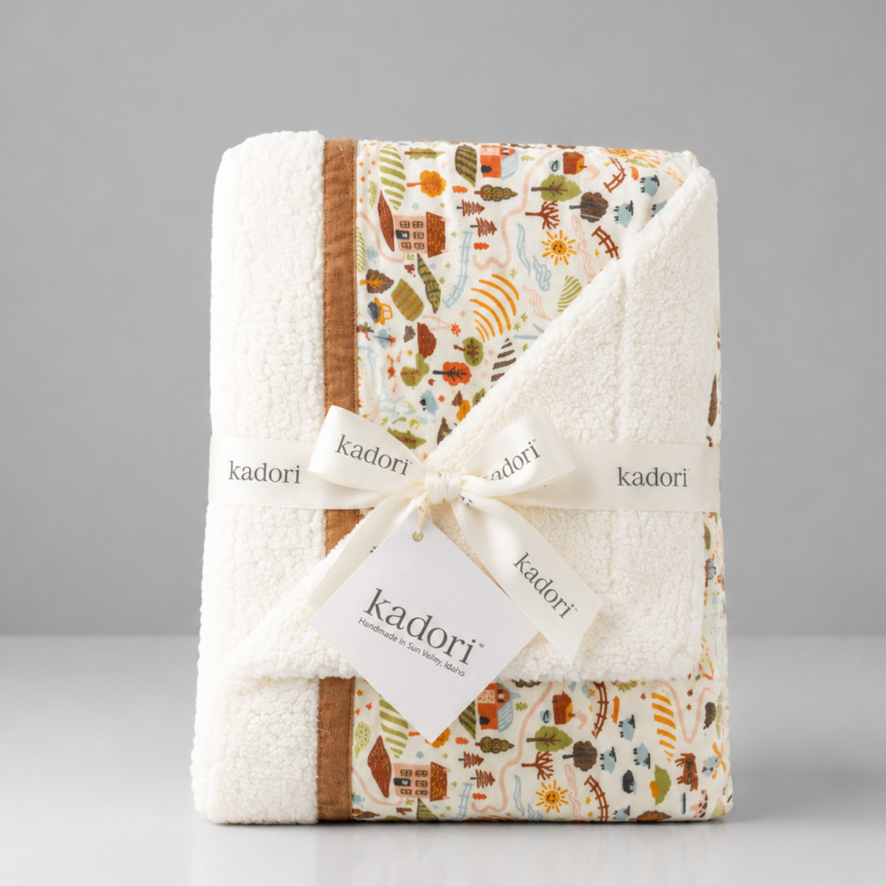 A folded Kadori baby blanket with a cream sherpa border and a multicolor autumn-themed patchwork pattern, tied with a white ribbon and tag.