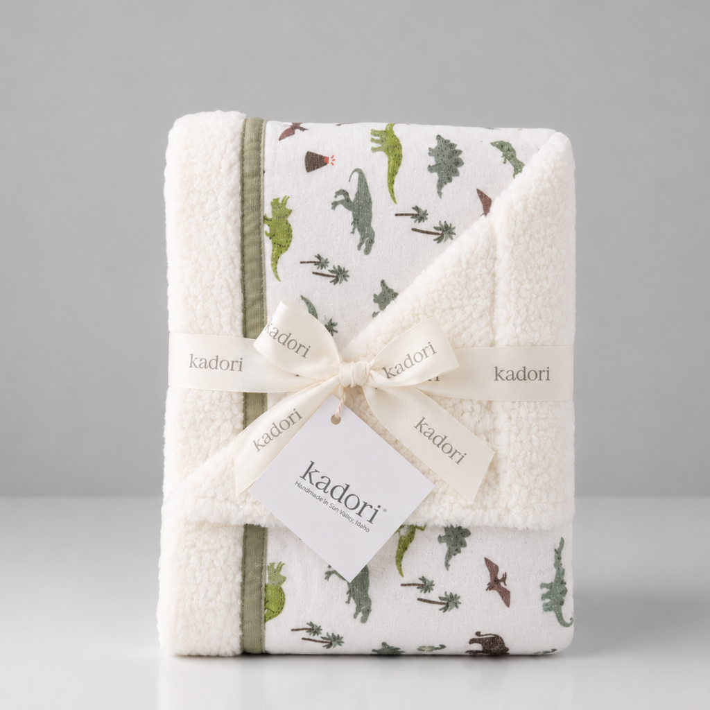 A folded white plush baby blanket with a green dinosaur print and olive green trim, wrapped in a cream ribbon with a Kadori tag, on a light gray background.