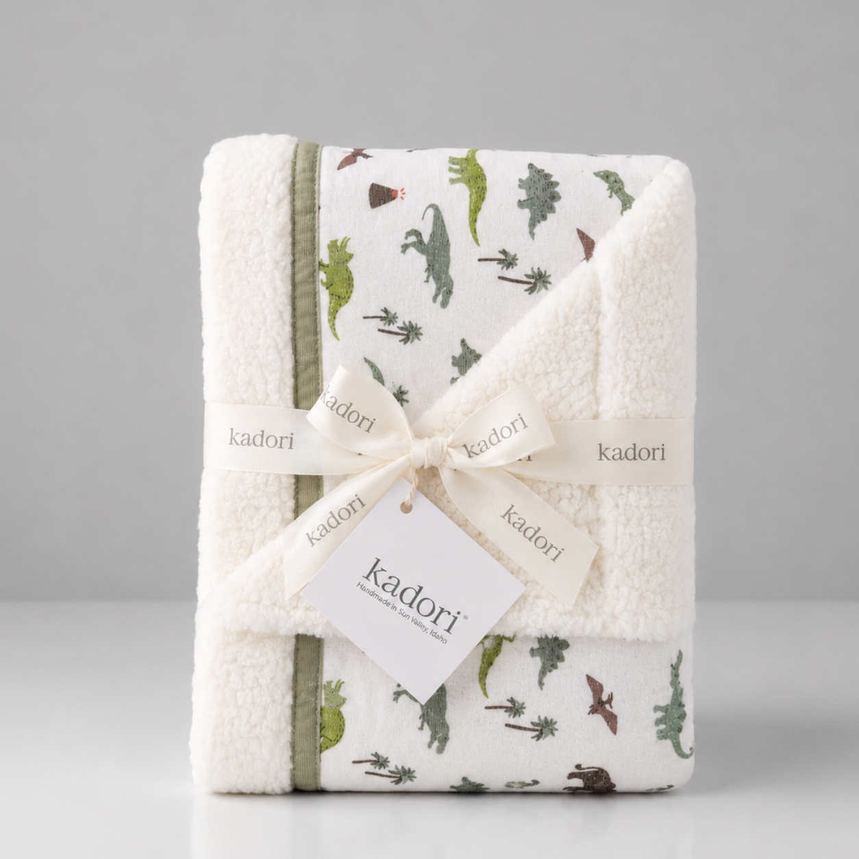 A folded white plush baby blanket with a green dinosaur print and olive green trim, wrapped in a cream ribbon with a Kadori tag, on a light gray background.