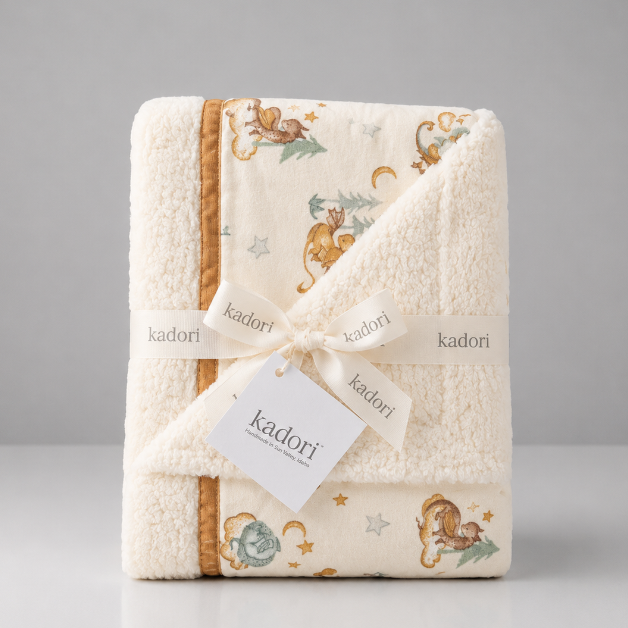 A folded plush baby blanket with a hood, featuring a cream background with a safari print of monkeys, stars, and moons, tied with a white ribbon printed with the brand name 'kadori'.
