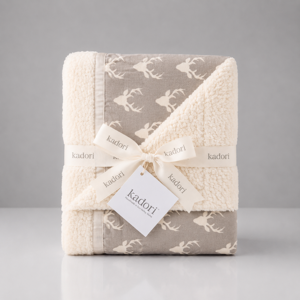 A folded Kadori baby blanket with a soft sherpa fleece texture, featuring a taupe-gray and cream deer pattern, wrapped in a cream ribbon with a Kadori tag.